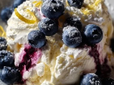 A scoop of blueberry lemon cheesecake ice cream in a cone topped with fresh blueberries.