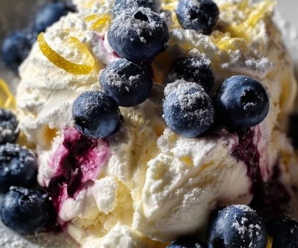A scoop of blueberry lemon cheesecake ice cream in a cone topped with fresh blueberries.