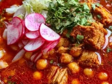 A bowl of flavorful red pozole garnished with fresh toppings and spices.