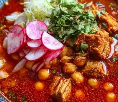 A bowl of flavorful red pozole garnished with fresh toppings and spices.