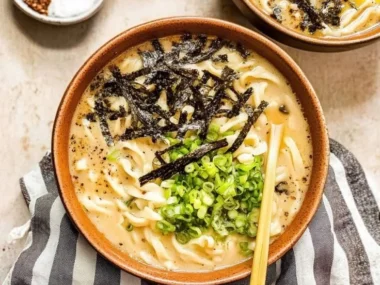 A steaming bowl of udon noodle soup with vibrant vegetables and tofu on a wooden table